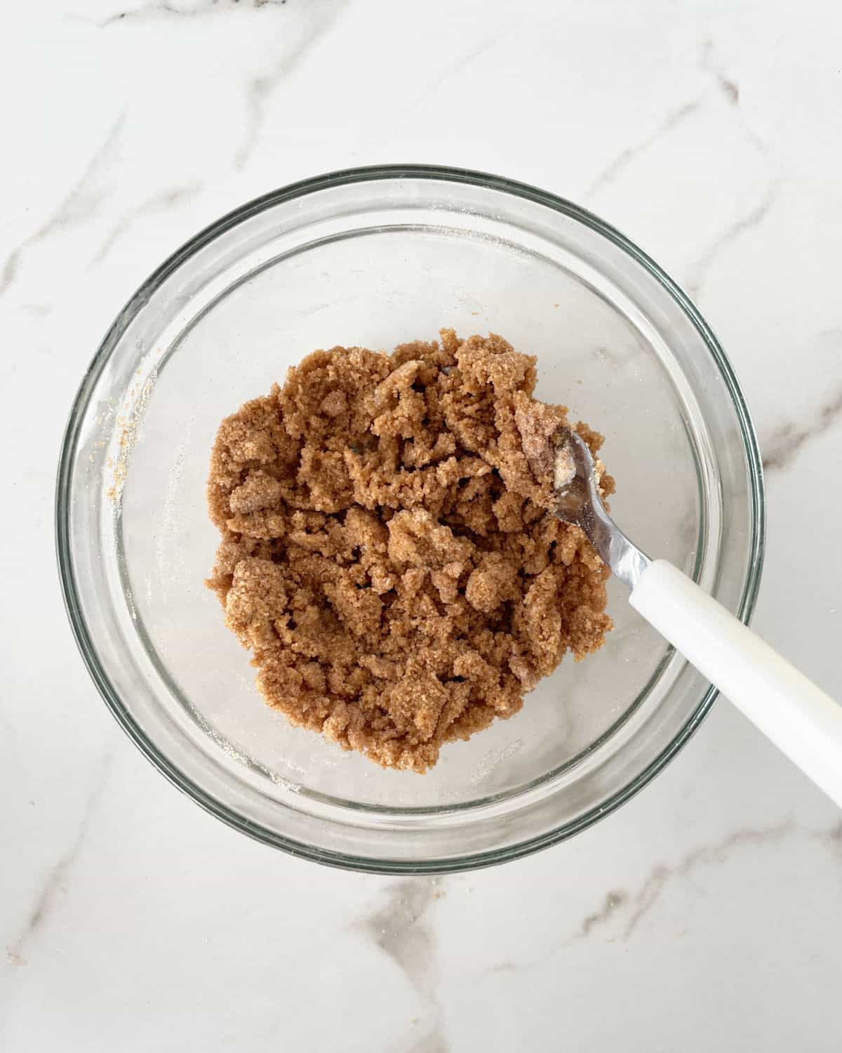 Mixed brown sugar cinnamon streusel in a glass bowl with white spoon in it. White marble surface.