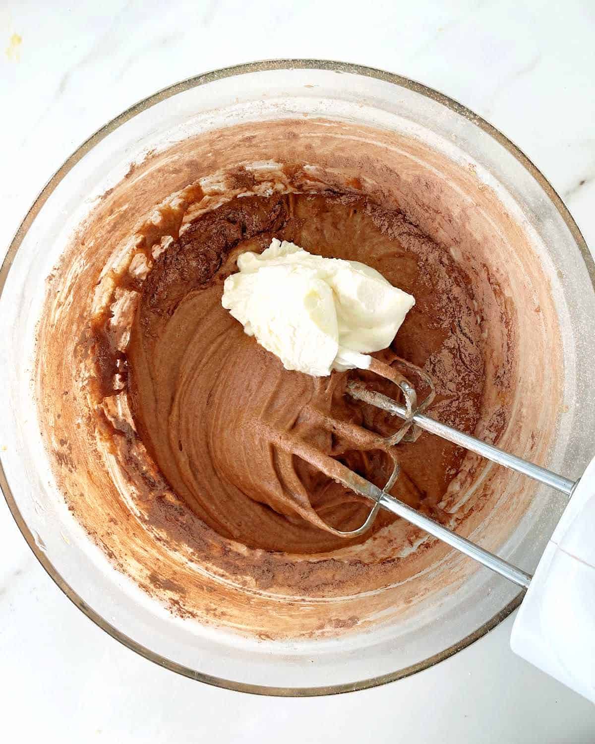 Sour cream added to chocolate cake batter in a glass bowl with electric beaters in it. White surface.