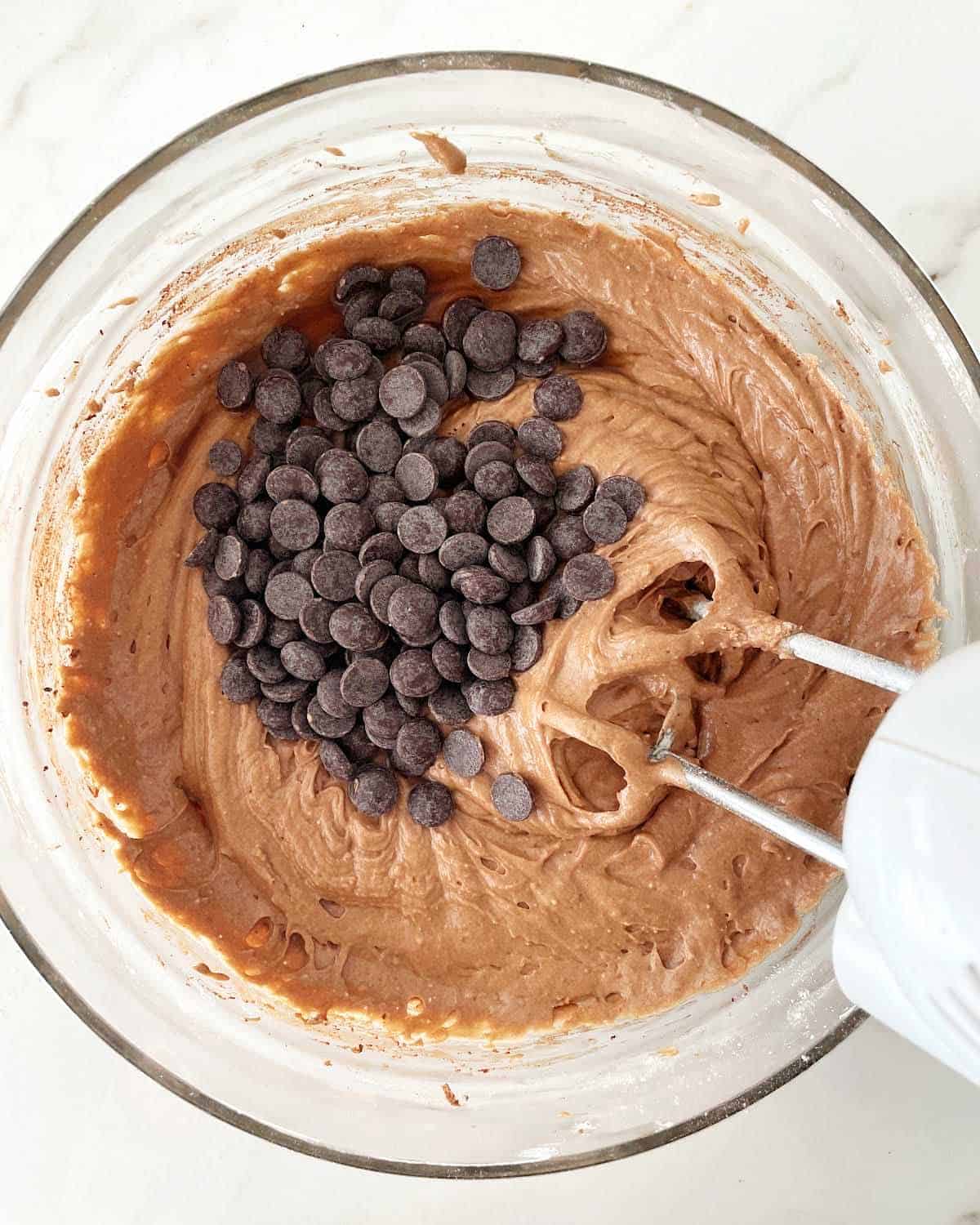 Chocolate chips added to chocolate cake batter in a glass bowl with an electric mixer inside. White marble surface.