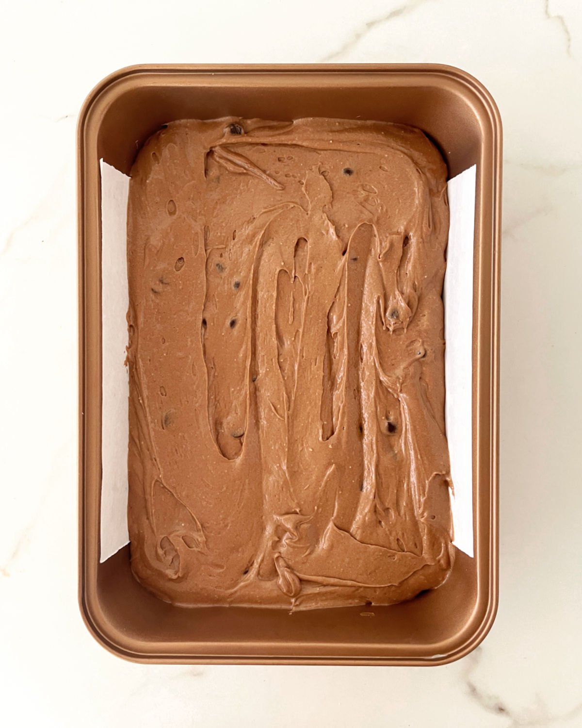 Top view of chocolate cake batter in a rectangular copper colored pan on a white marble surface.