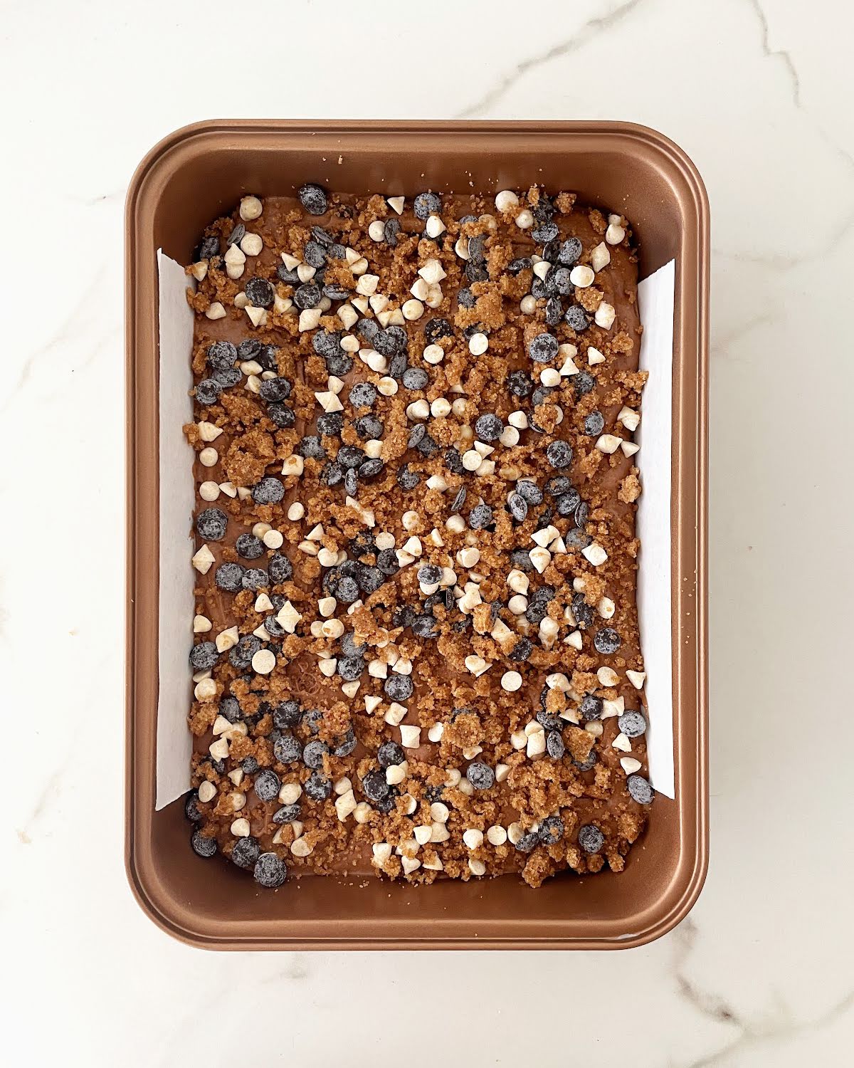 Different colors of chocolate chips topping a chocolate cake batter in a copper pan on a white marble surface.