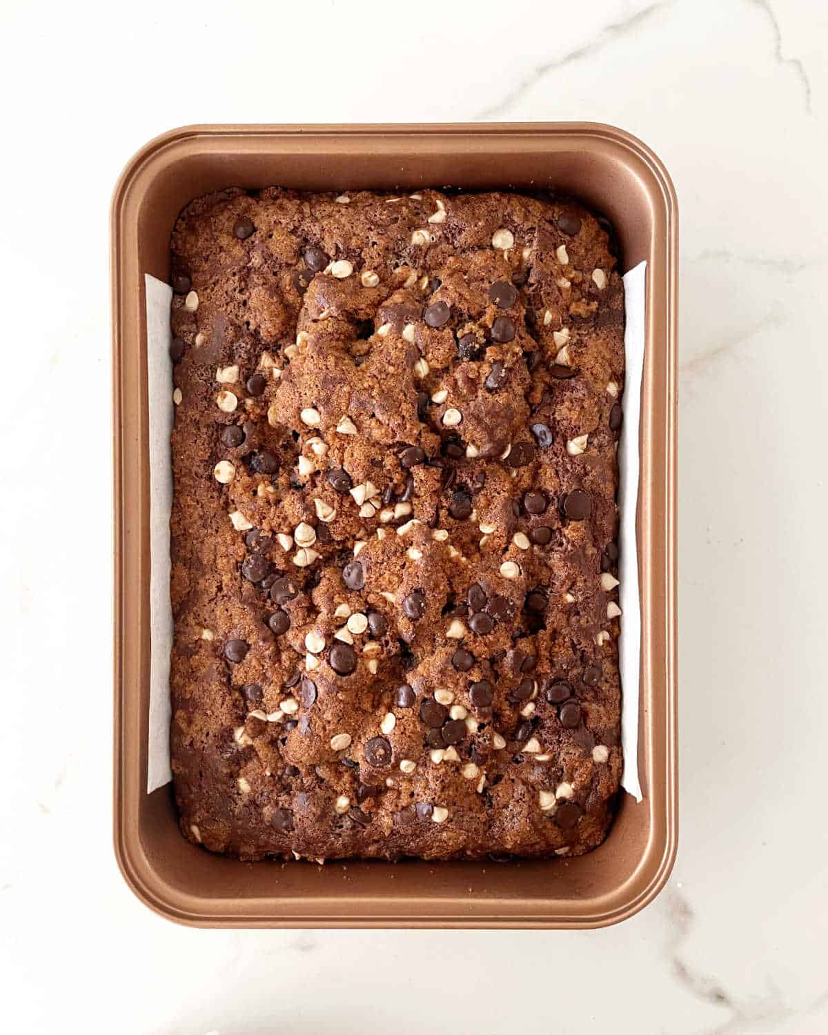 Baked chocolate cake topped with chocolate chips in a copper colored pan on a white marbled surface.