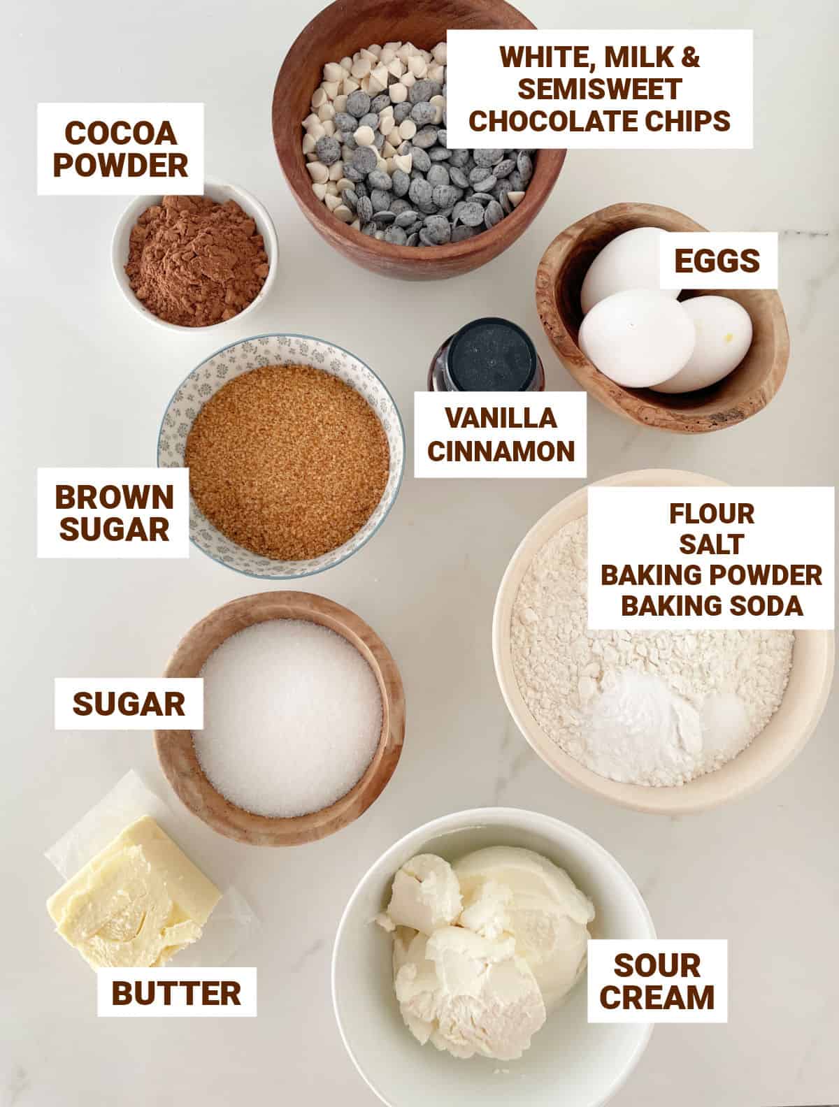 White marble with bowl containing ingredients for chocolate cake including chips, sugars, butter, eggs, flour, sour cream, vanilla, flour mixture.