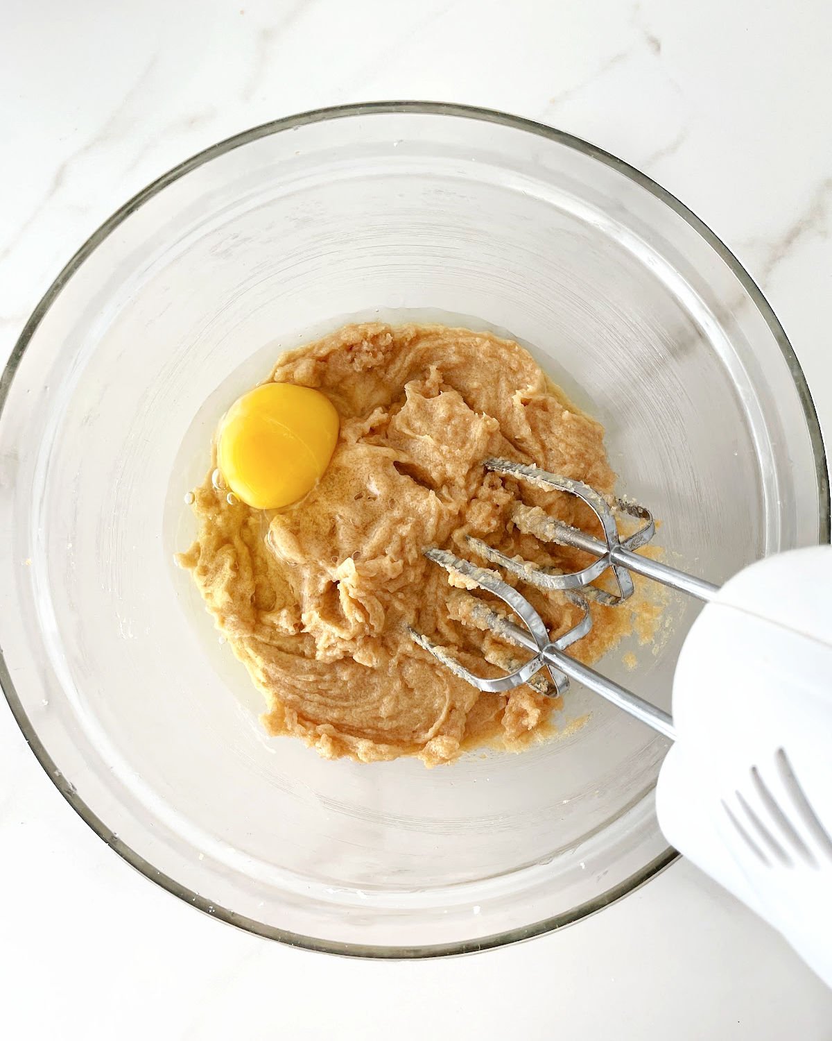 Egg added to sugar butter mixture in a glass bowl on a white surface with an electric mixer inside.
