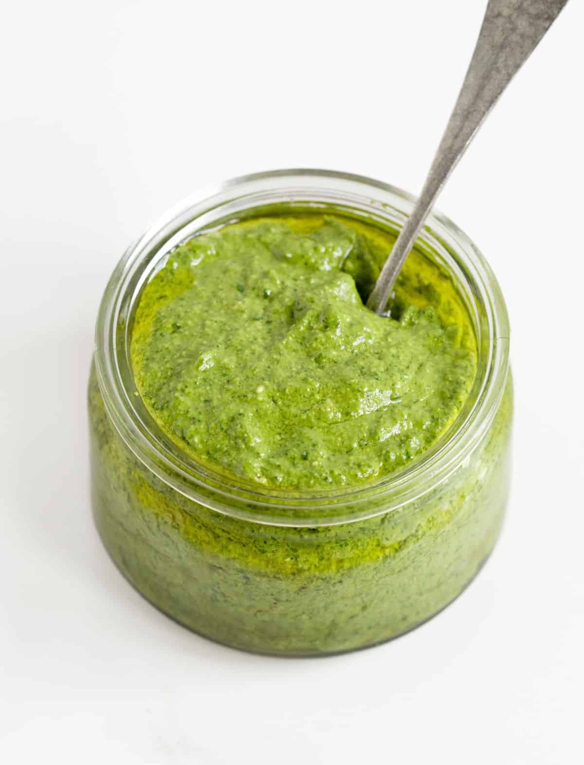 Jar of spinach pesto with a vintage silver spoon on a white surface. 