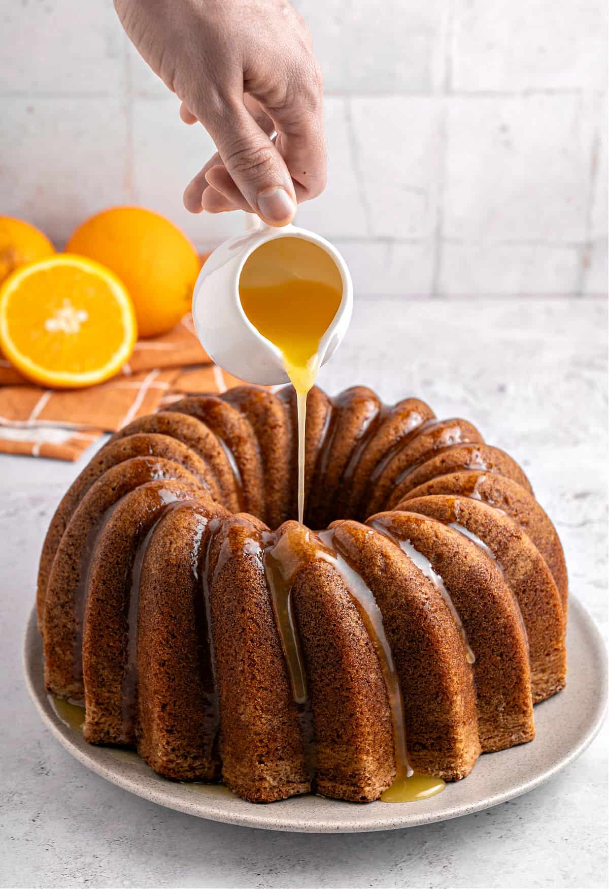 Pouring orange glaze on a bundt cake. White surface and background. Fresh oranges.