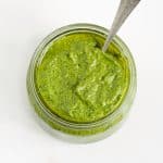 Top view of jar with spinach pesto and a vintage silver spoon inside. White surface.