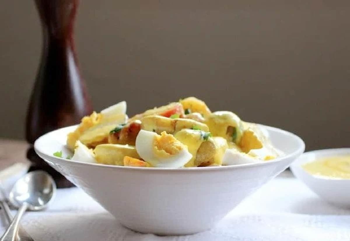 White bowl of Egg Potato Salad, white cloth, spoon, sauce, pepper shaker.