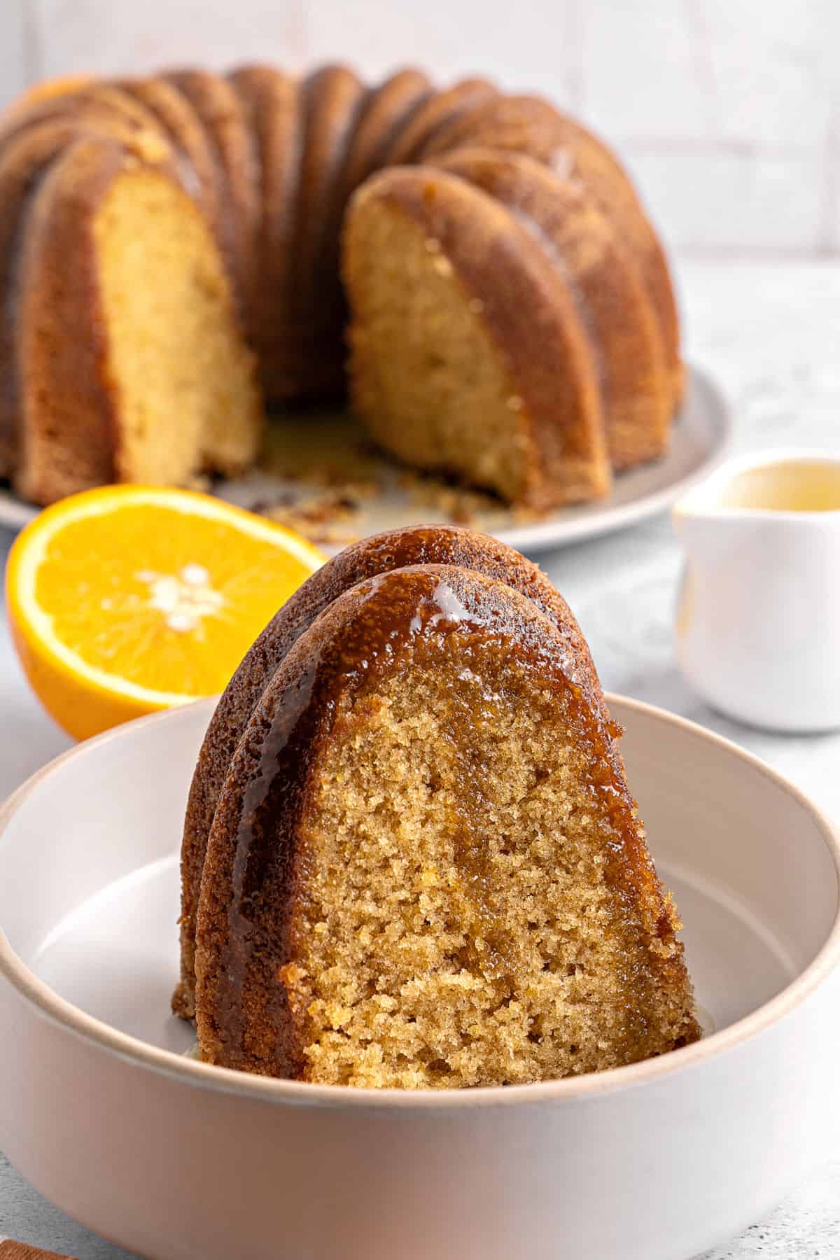 Slice of glazed orange bourbon bundt cake in a bowl. Whole cake in the white background.