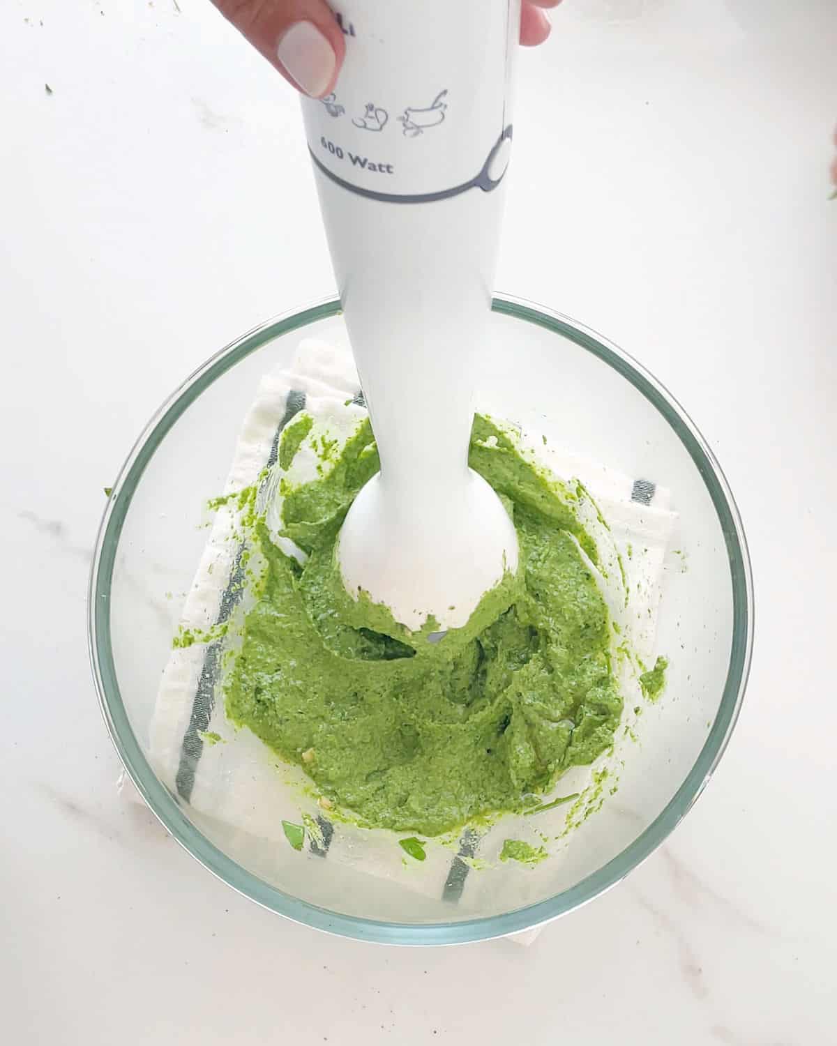 Mixing pesto with an immersion blender in a glass bowl on a white surface.