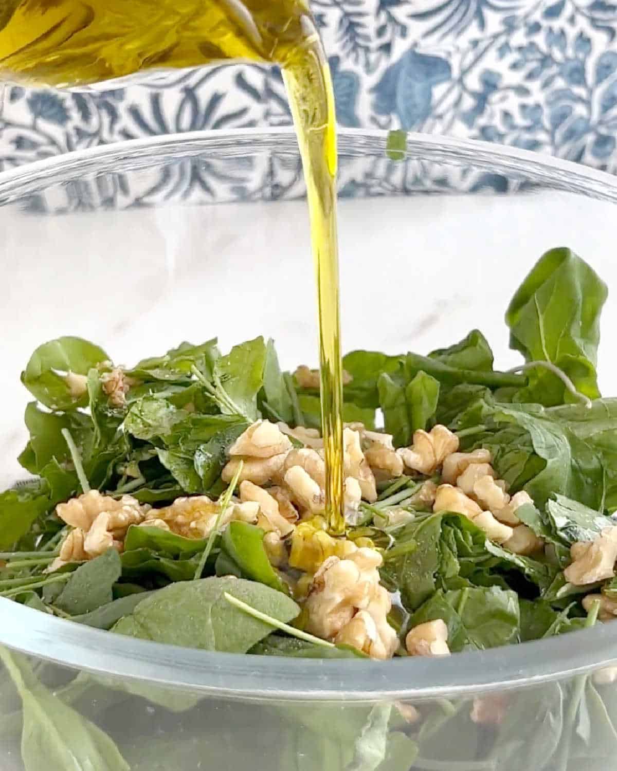 Adding oil to a glass bowl with walnuts and spinach leaves.