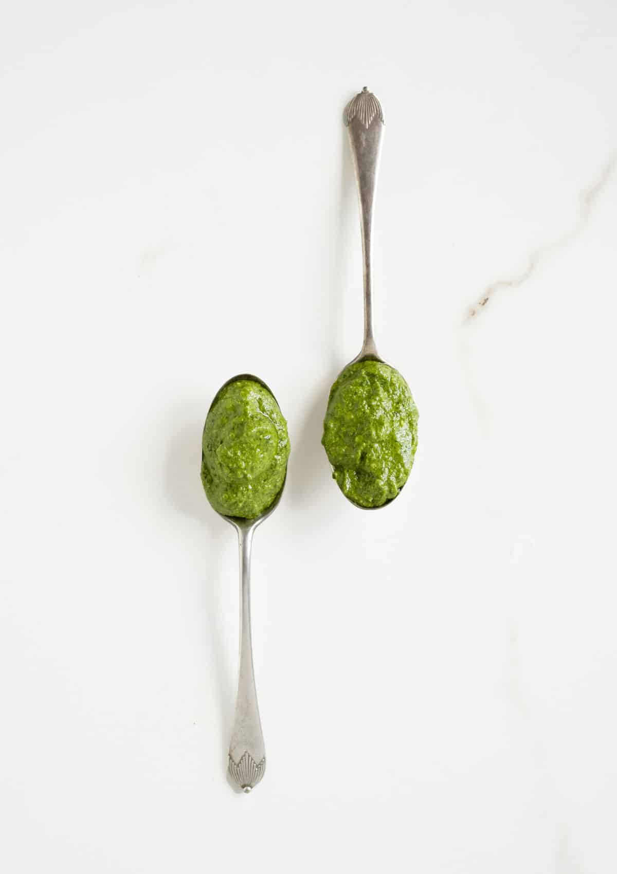 Two vintage spoons with spinach pesto on a white marble surface.