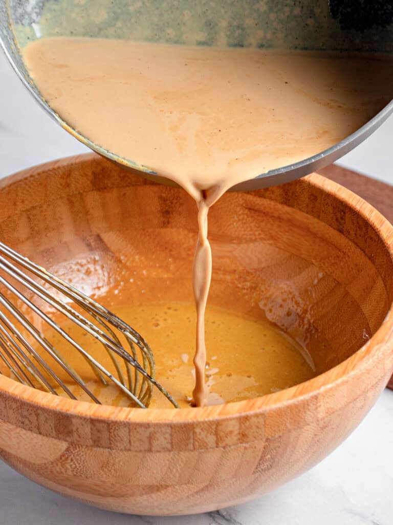 Pouring coffee cream into beaten egg yolks in a wooden bowl. Wire whisk.