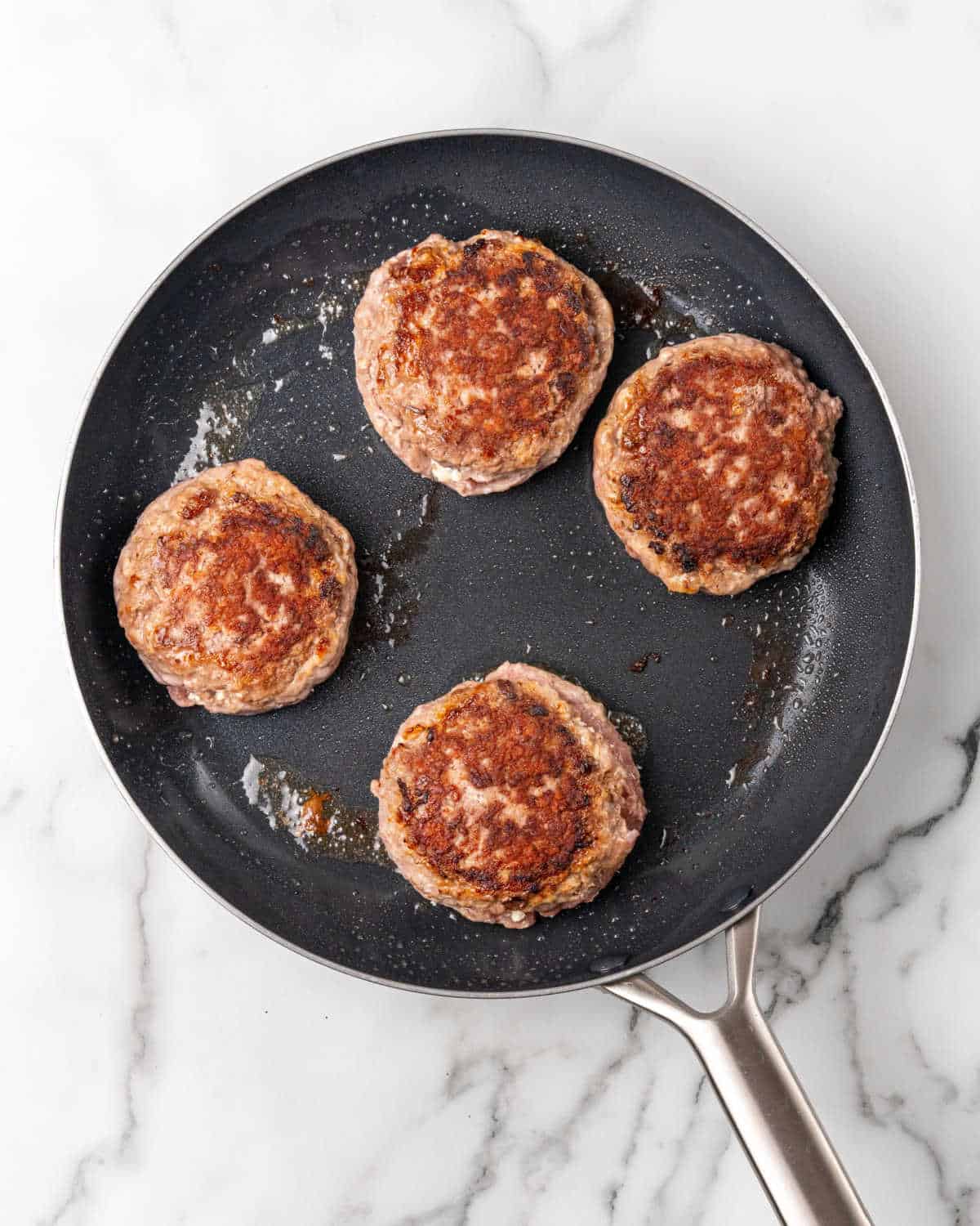 Four hamburgers being cooked in a dark skillet. White and grey marble surface.