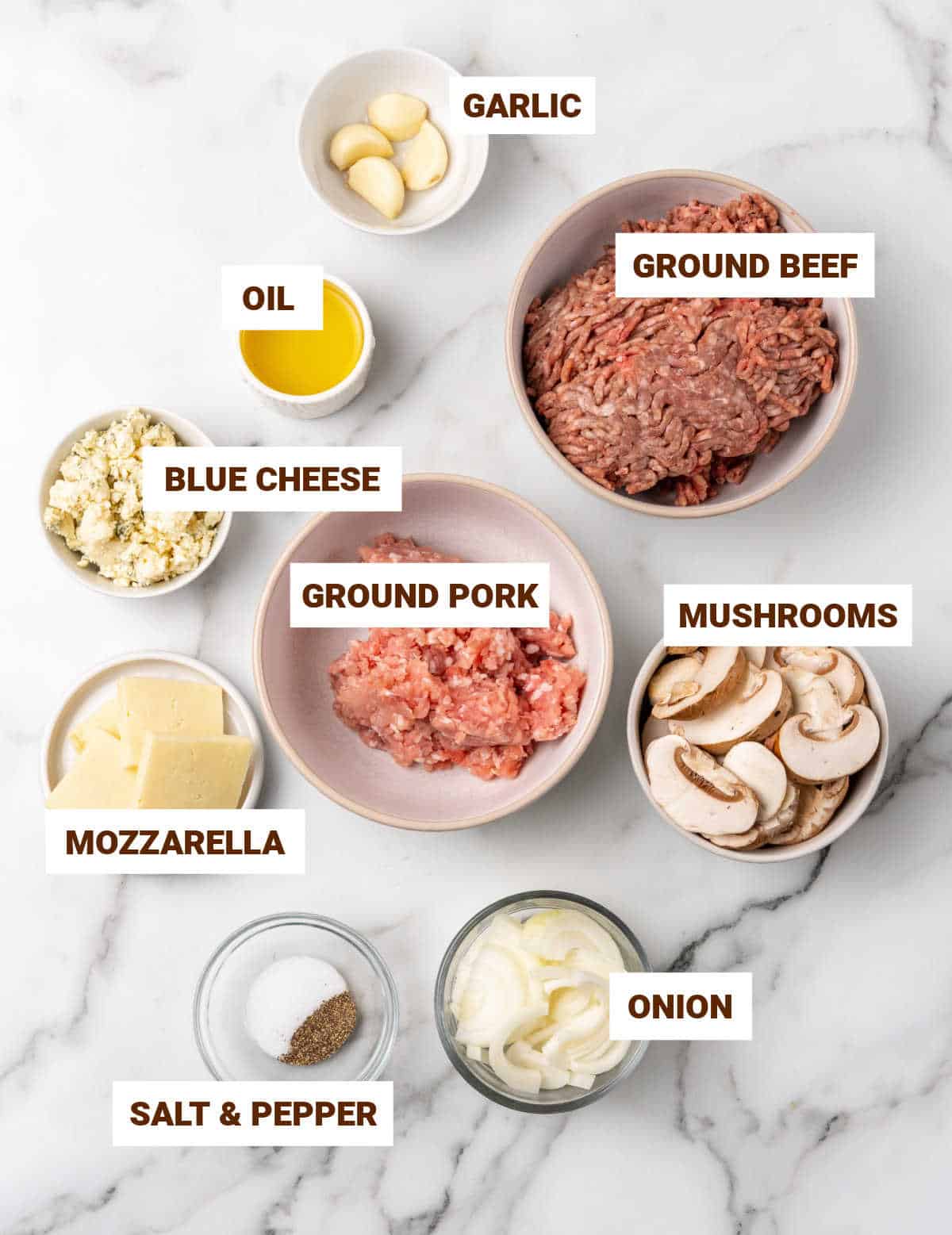 White marble surface with bowls containing ingredients for blue cheese burgers including mushrooms, garlic, cheeses, seasonings, ground meats.