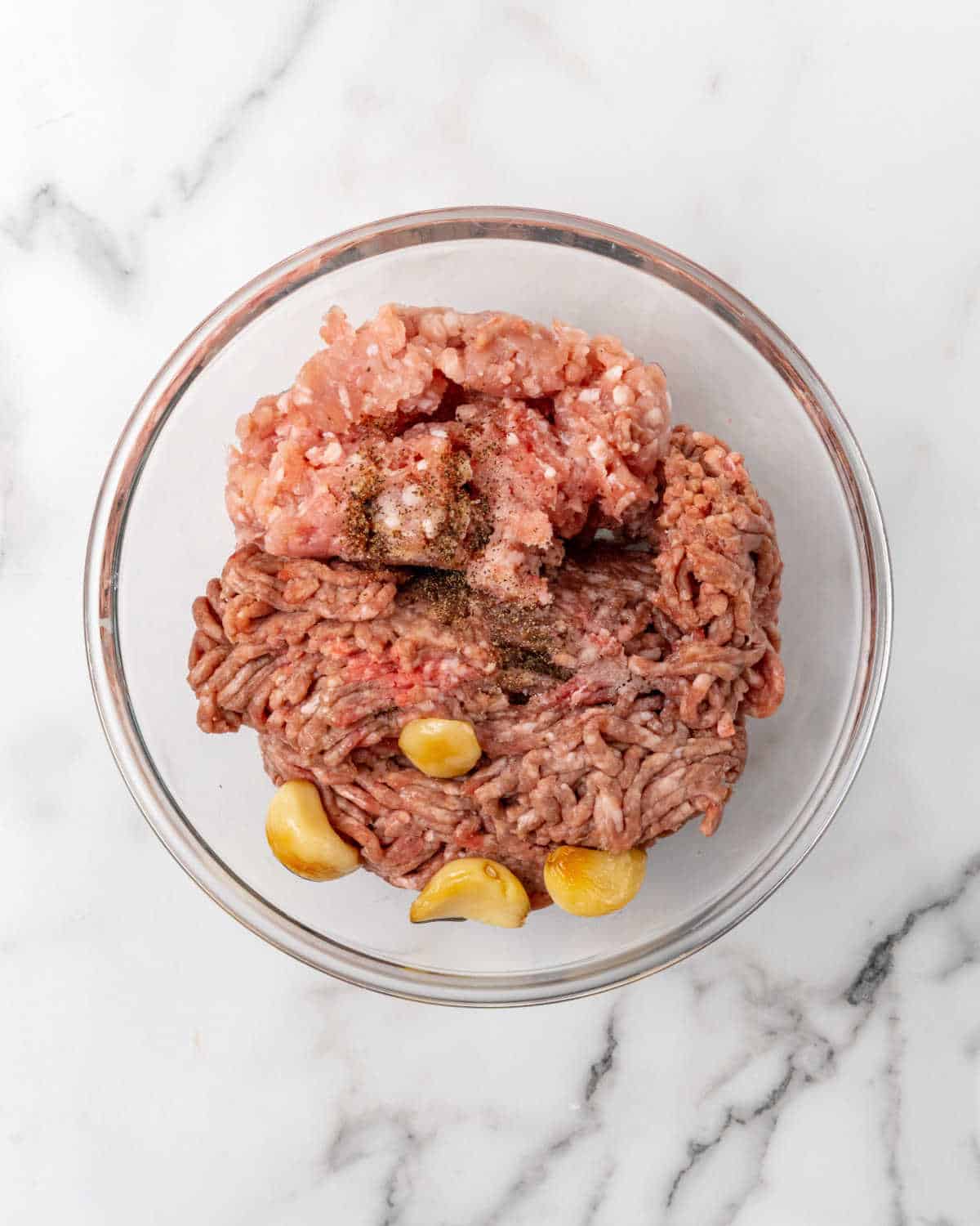 Glass bowl with ground beef, garlic cloves and spices on a white marble surface.