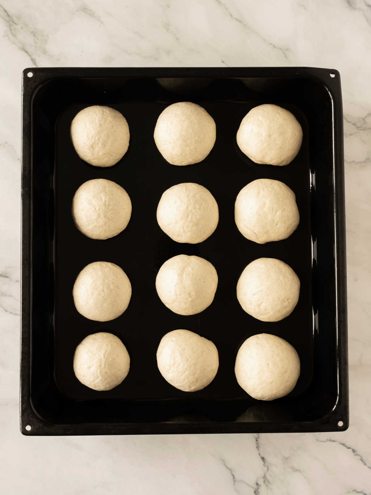 Three rows of bread dough balls on a black oven tray. White marbled surface.