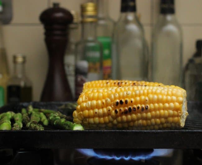 Charring corn and asparagus on black skillet over a flame; empty bottles and pepper mill in background