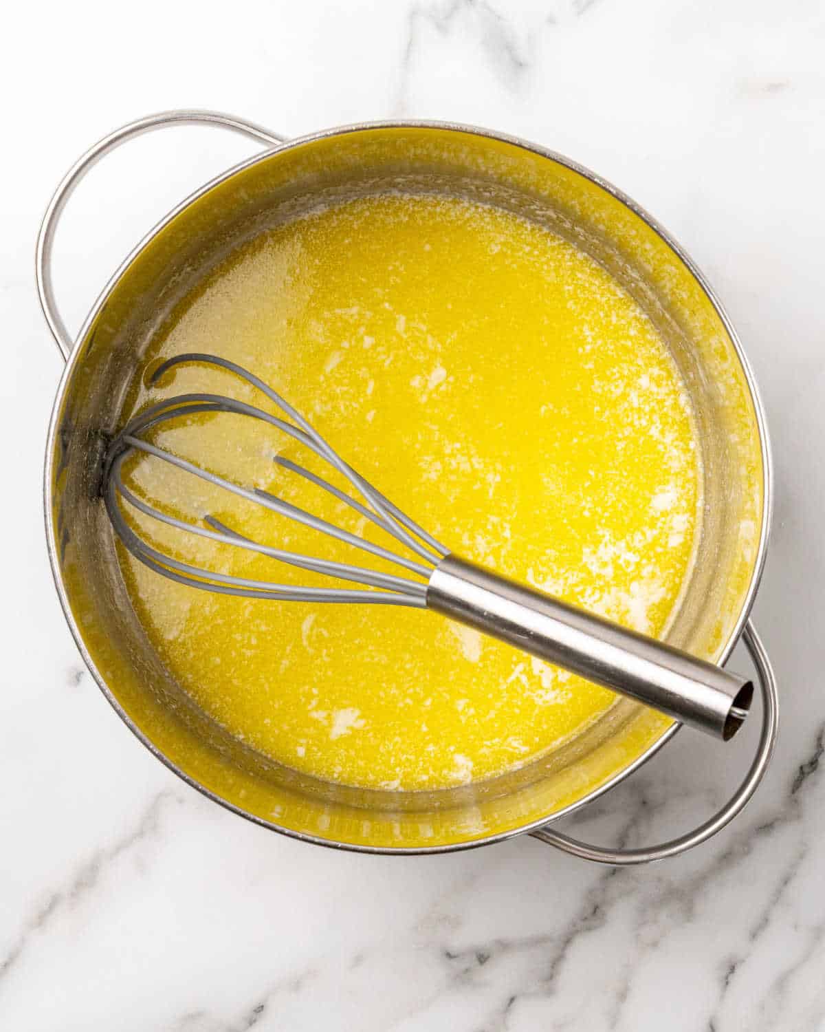 Large metal saucepan with passionfruit curd and a whisk on white marble with grey stripes.