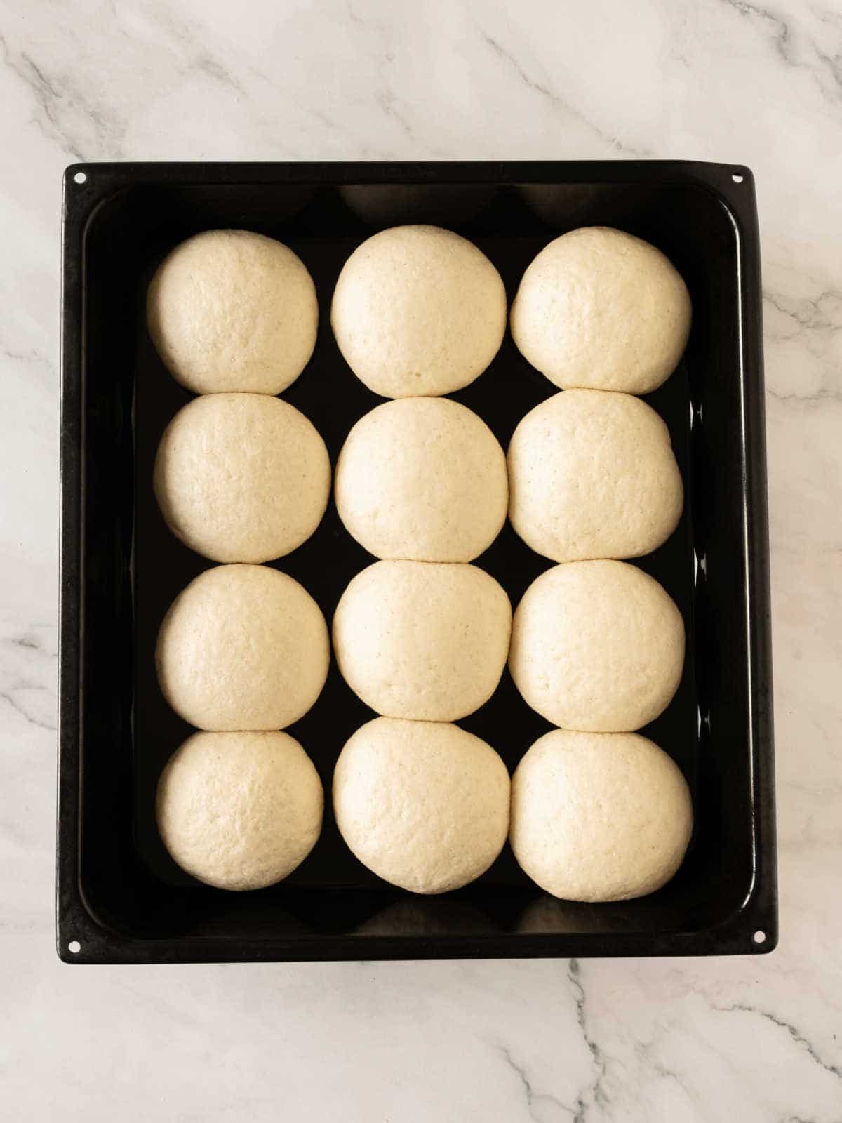 Puffed bread dough balls in rows on a black oven tray. White marbled surface.