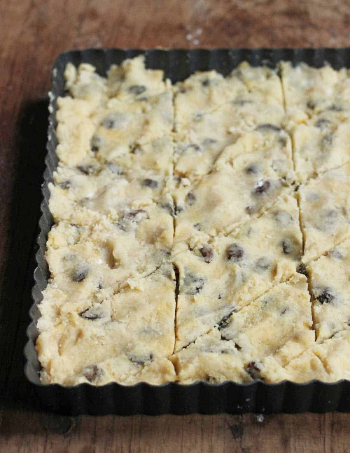 Scored unbaked raisin shortbread dough in a dark metal square pan on a wooden table.