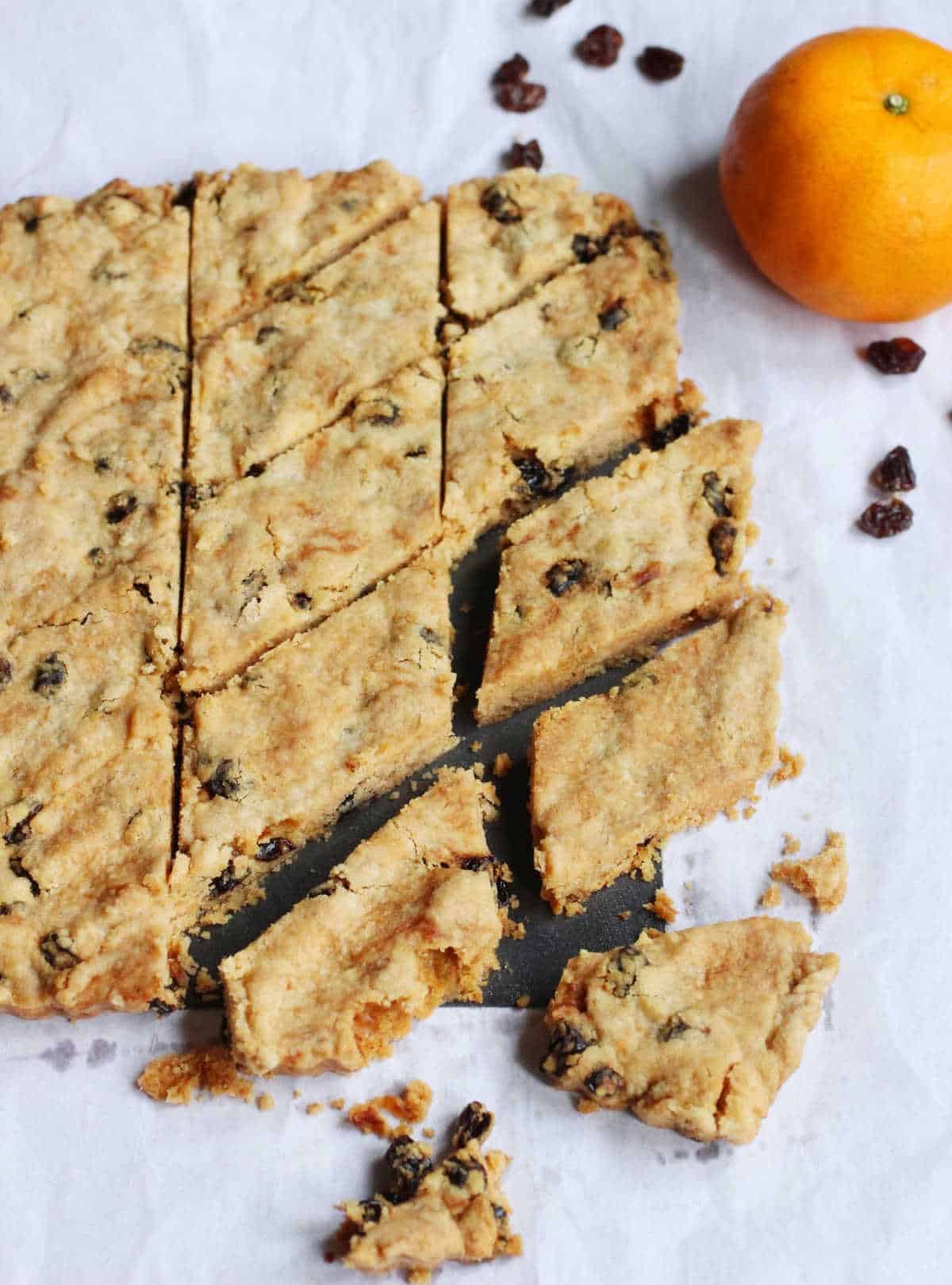 Squares of raisin shortbread on a white piece of paper. An orange and loose raisins around. 