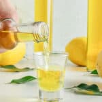 Close up image of limoncello shot being poured. Grey background, liqueur bottles.