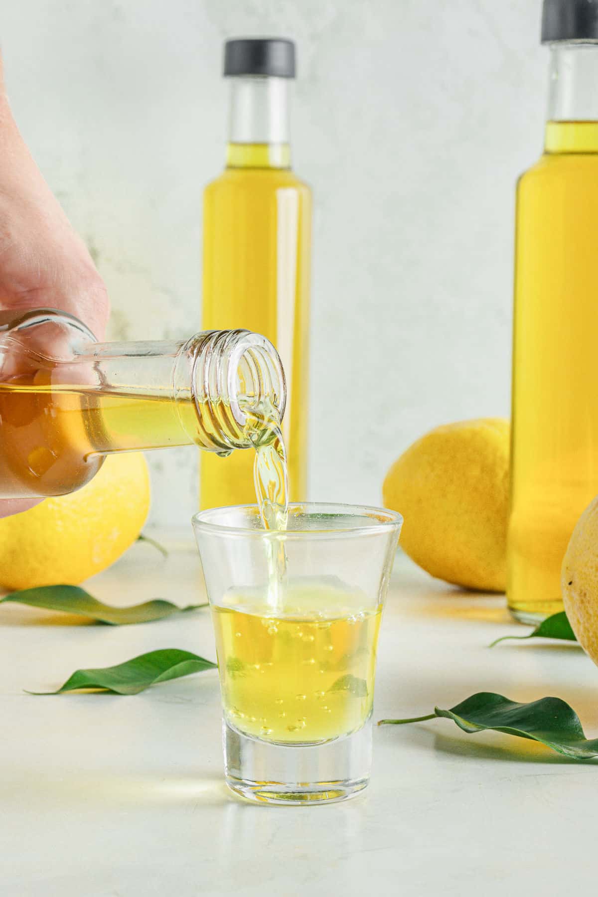 Lemon liqueur bottles, gray background, lemons and hand serving a shot glass of limoncello.