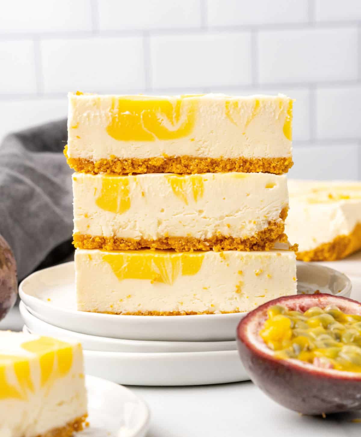 On a stack of white plates, three squares of cheesecake with passion fruit are stacked. White tile background, fresh half fruit, blue cloth. 