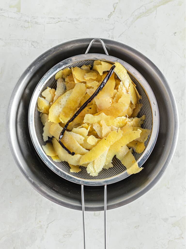 Straining lemon vodka with vanilla bean on a metal colander over a metal bowl.