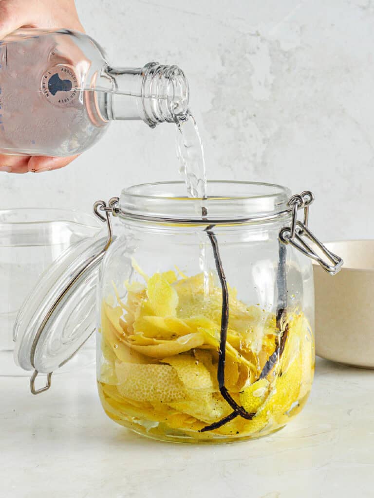 Pouring vodka into a mason jar with lemon peel and vanilla bean. Gray background. 