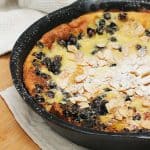 Cast iron skillet with partial view of blueberry white chocolate pancake. Beige cloth and wooden board below.