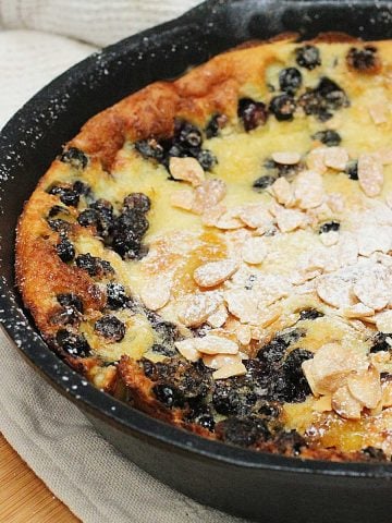 Cast iron skillet with partial view of blueberry white chocolate pancake. Beige cloth and wooden board below.