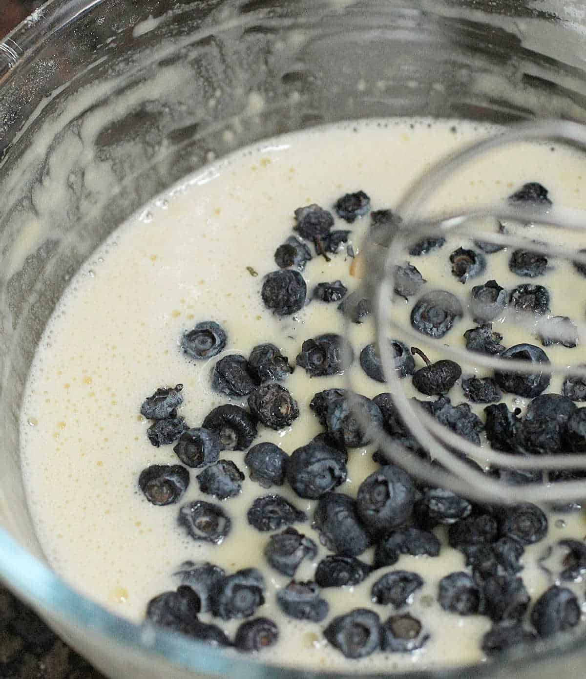 Glass bowl with pancake batter and blueberries and a whisk.