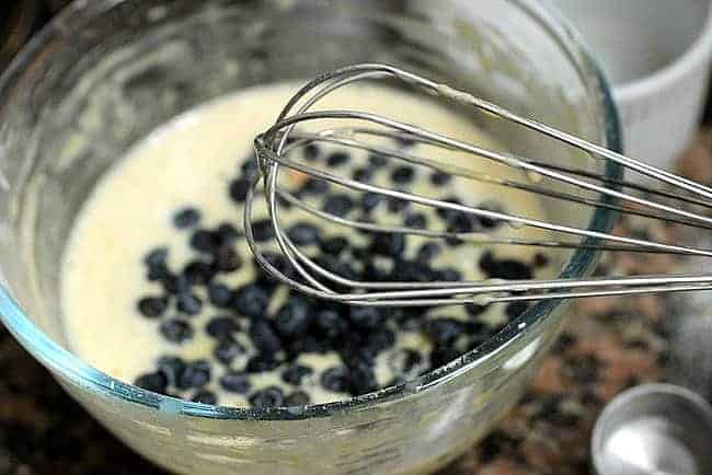 Glass bowl with pancake batter and blueberries and a whisk.