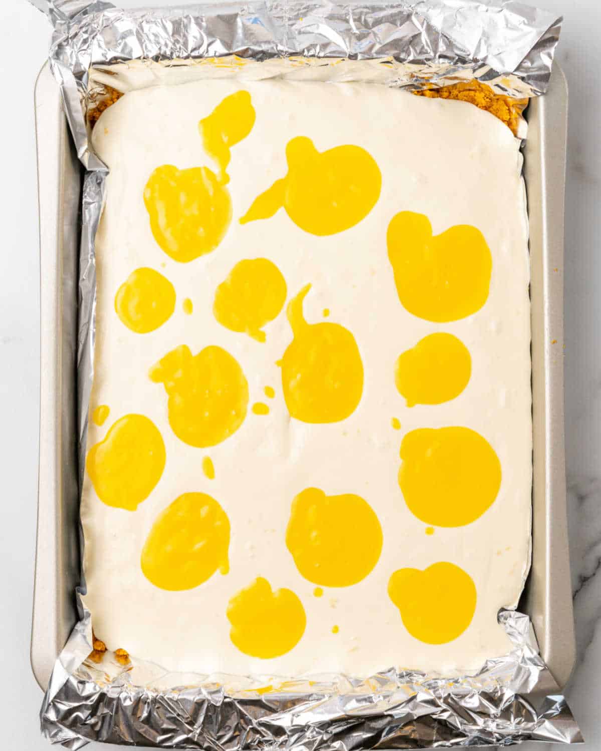 No bake cheesecake batter with mounds of passionfruit curd in a foil-lined rectangular pan on a white surface.