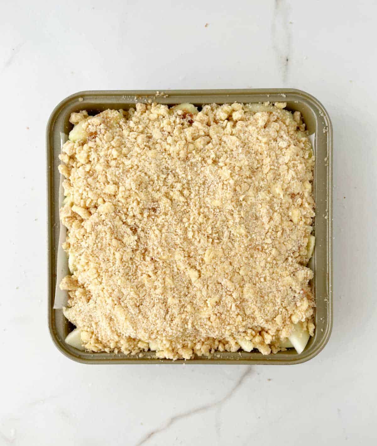 Crumble topping covering apples in a square baking pan on a white marble surface.