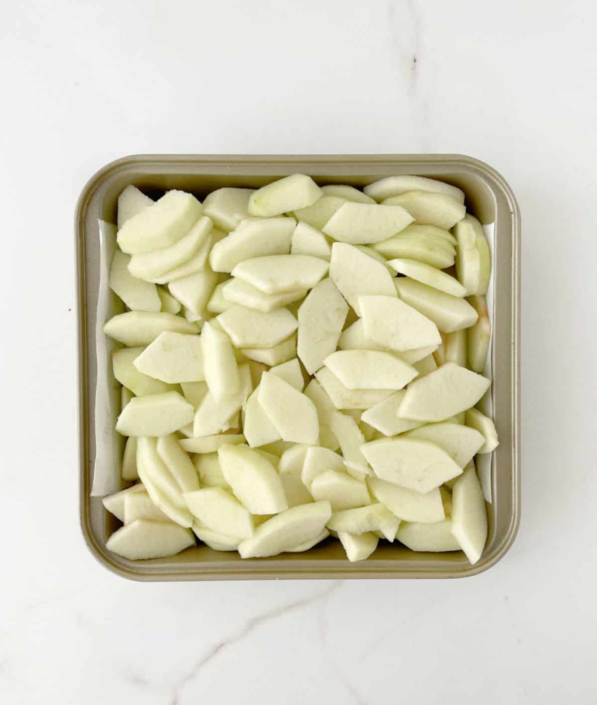 Sliced apples in a square baking pan on a white marble surface. 