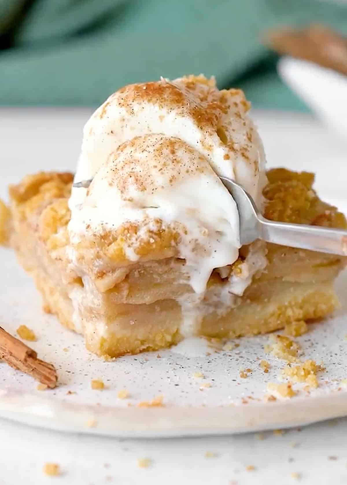 Fork cutting a square of apple crumb bar with ice cream on a white plate. Green background. 