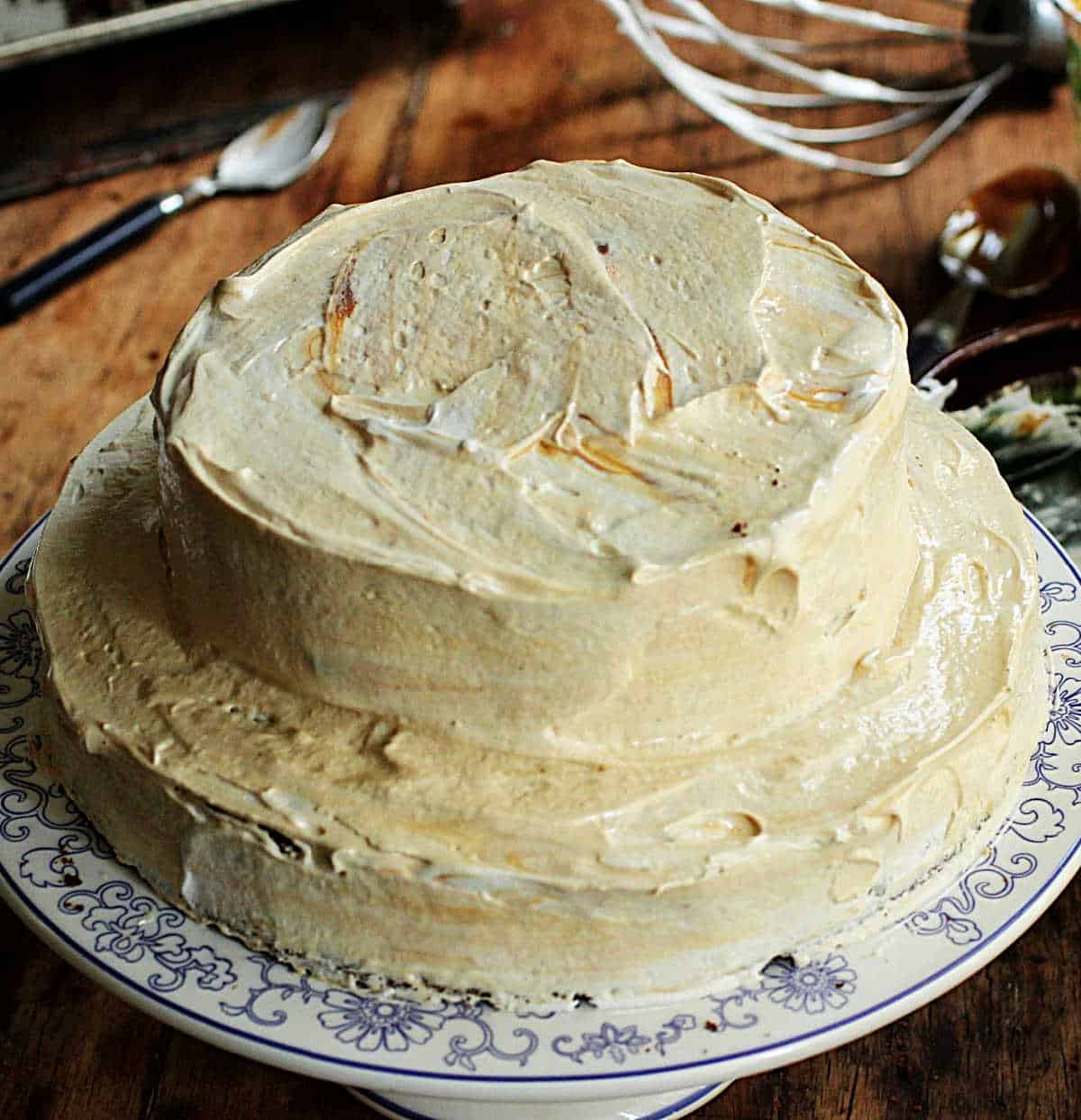 Whole meringue frosted cake on white blue cake stand, wooden table, utensils scattered around.