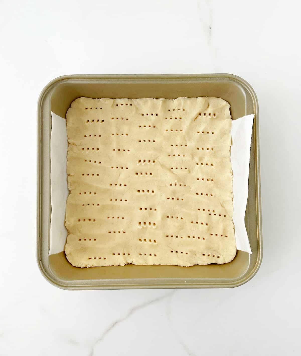 Parchment lined square baking pan on a white surface with pricked shortbread crust.