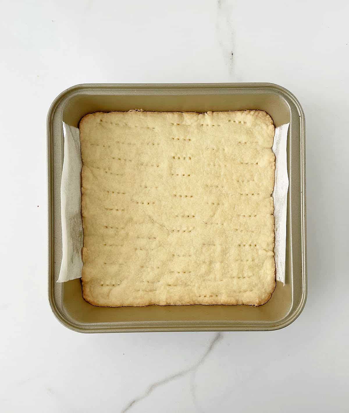 Pre baked shortbread crust on a parchment lined golden colored square pan on white marble surface. 