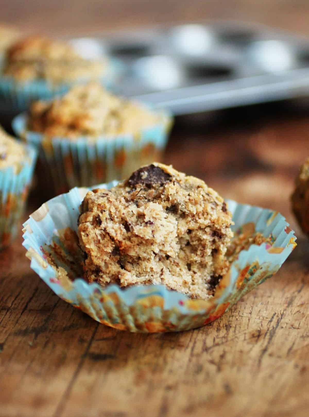 Half a banana chocolate chip muffin in opened blue paper liner on a wooden board. Pan and muffins in background. 