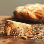 On a wooden table a slice of sweet bread with crunchy topping, scattered almonds, whole braid in background.