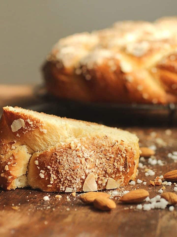 On a wooden table a slice of sweet bread with crunchy topping, scattered almonds, whole braid in background.