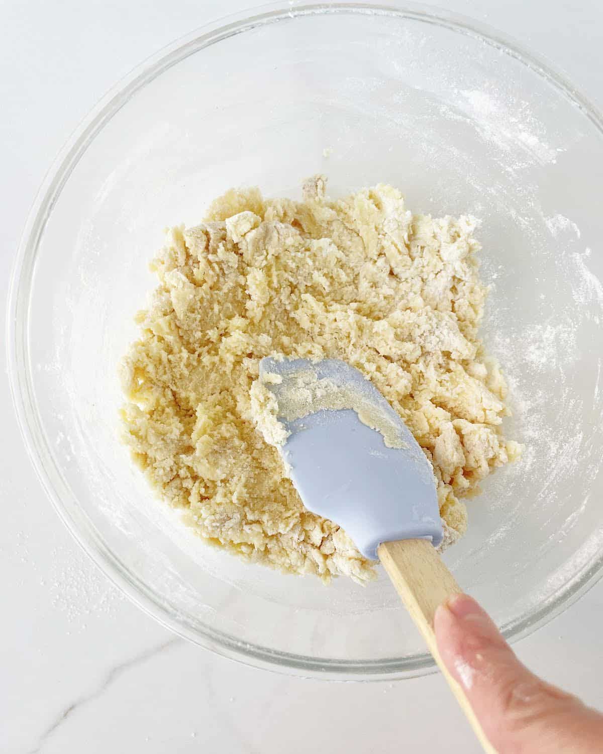 Mixing vanilla cookie dough in a glass bowl with a light blue spatula. White marble surface.