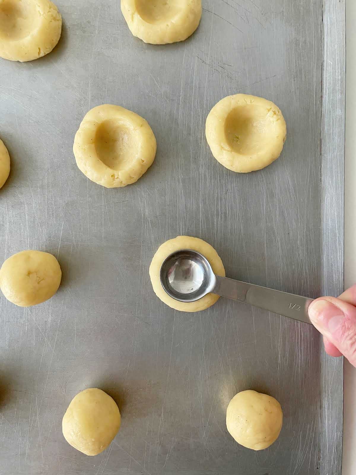 Metal cookie sheet with unfilled thumbprint cookies with hand making indentation with a spoon.