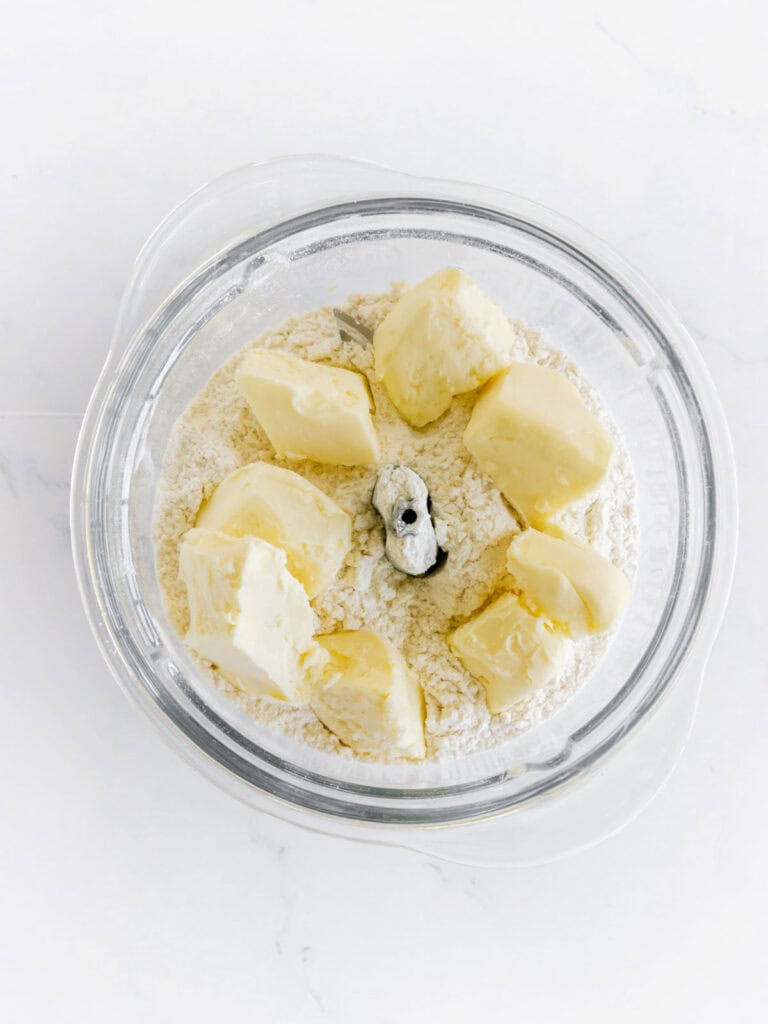 Butter pieces and dry ingredients in a food processor bowl on white marble.