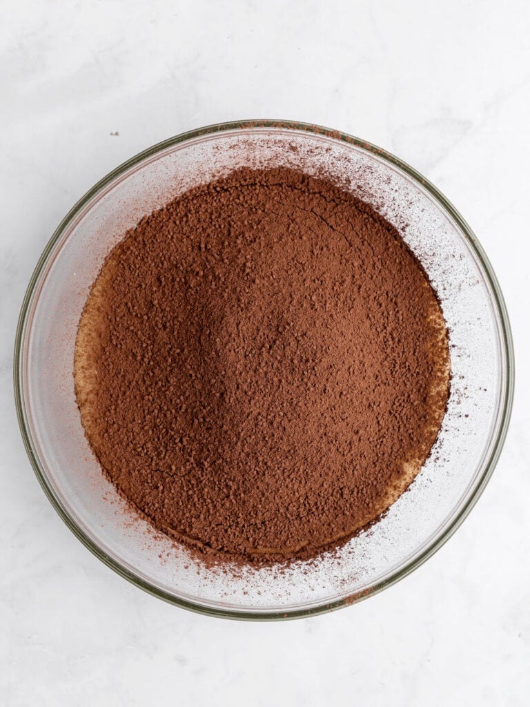 Sifted cocoa powder in a glass bowl on a white surface. Top view.