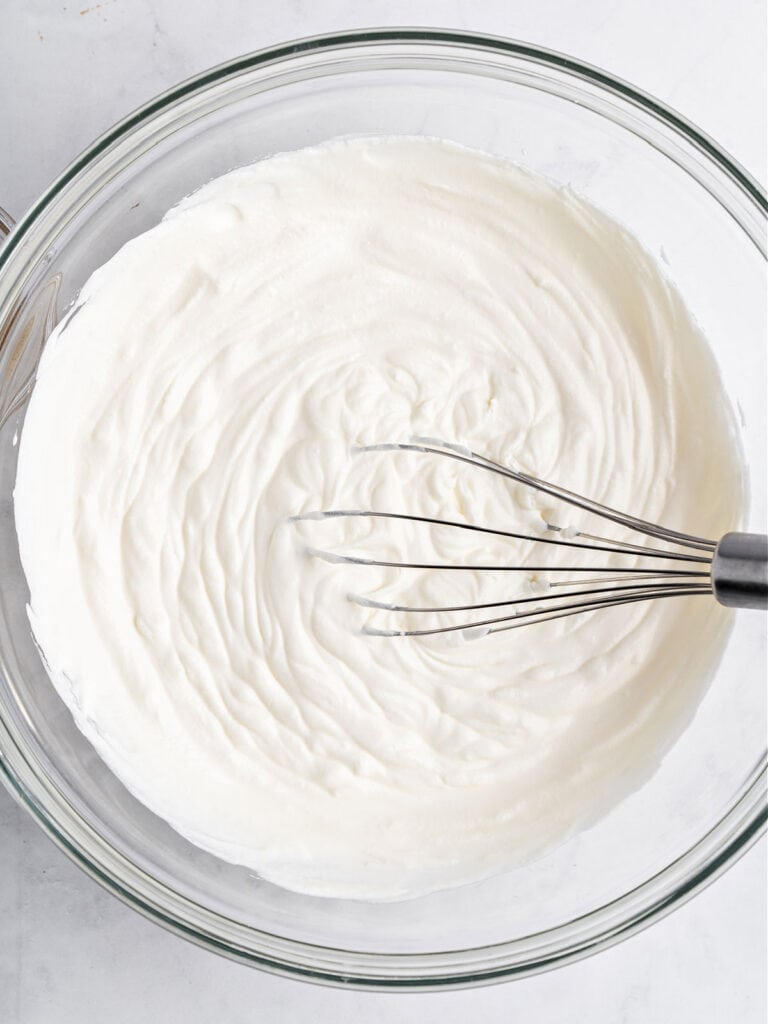 Whipped cream in a glass bowl with whisk. White surface. Top view.